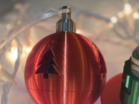 The red Christmas Money Puzzle Ornament, closed, with cut-out trees and a silver cap, in front of string lights.