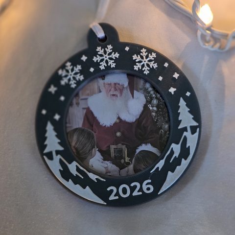 The ornament lying flat beside a string of lights, a white ribbon through the hanger.