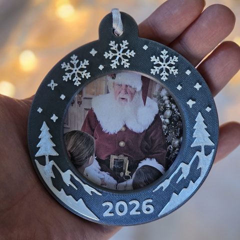 The Alpine Snow Photo Ornament in an open hand: snowflakes, pine trees and snowy mountains around a round photo window, with 2026 at the bottom.